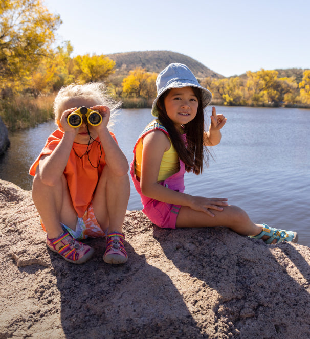 Two young girls wearing bright summer clothes and seacamp sandals sitting on rock in front of lake and looking through binoculars 