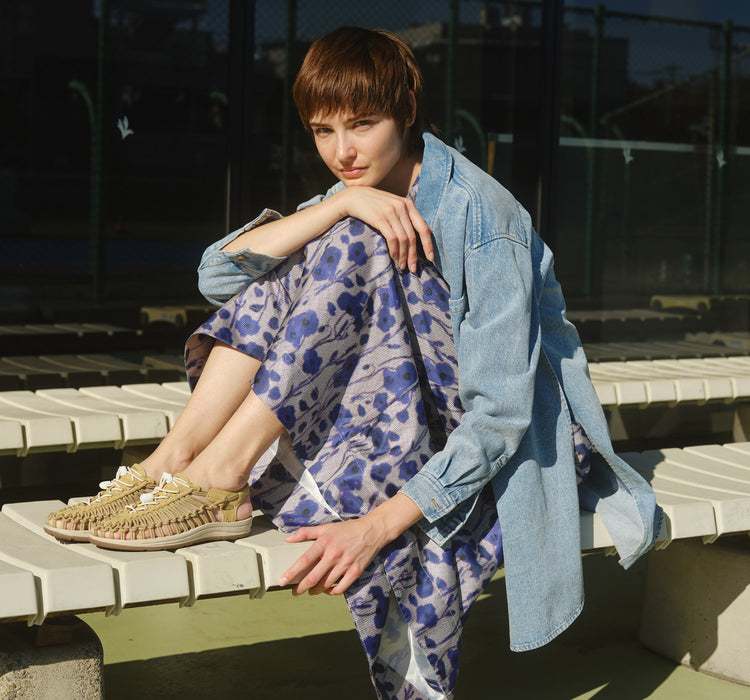 Woman wearing tan Uneeks and a blue, floral dress sitting on a white bench with her feet up. 