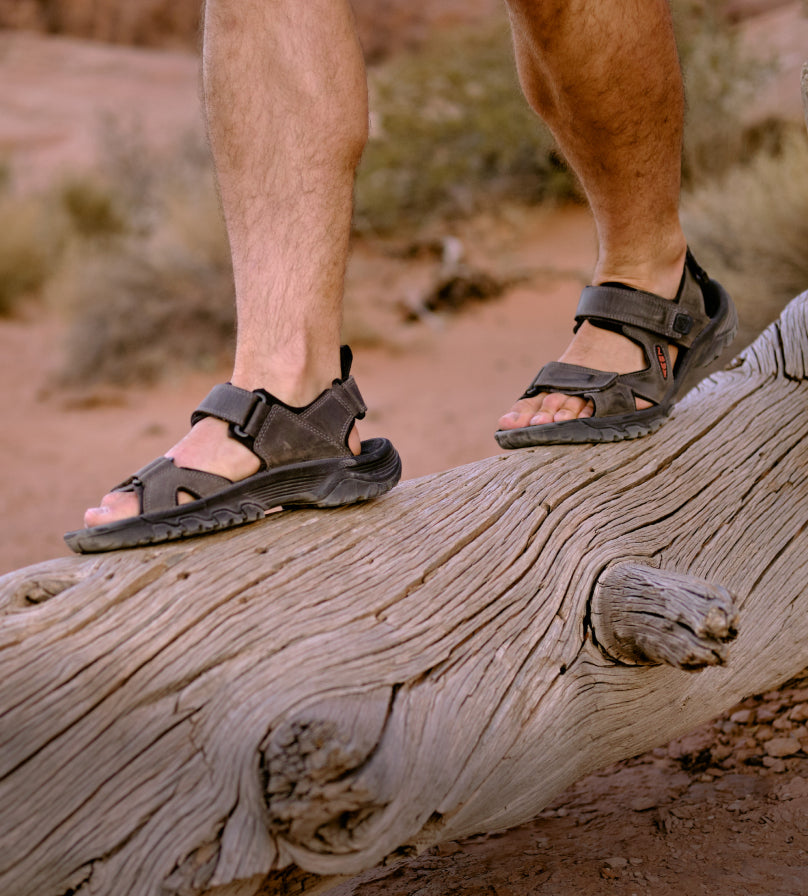 Knee-down shot of wearing Targhee III open-toe sandals and walking across a fallen tree on red dirt. 