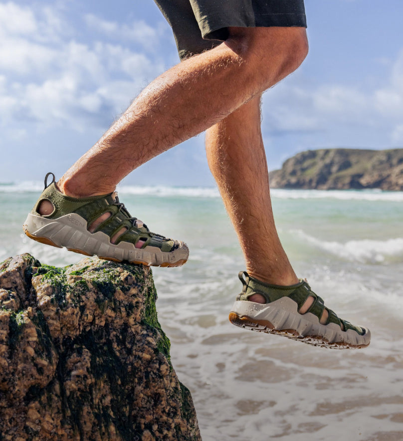 Knee-down shot of man wearing green and white Hyperport sandal and stepping of rock into the ocean. 
