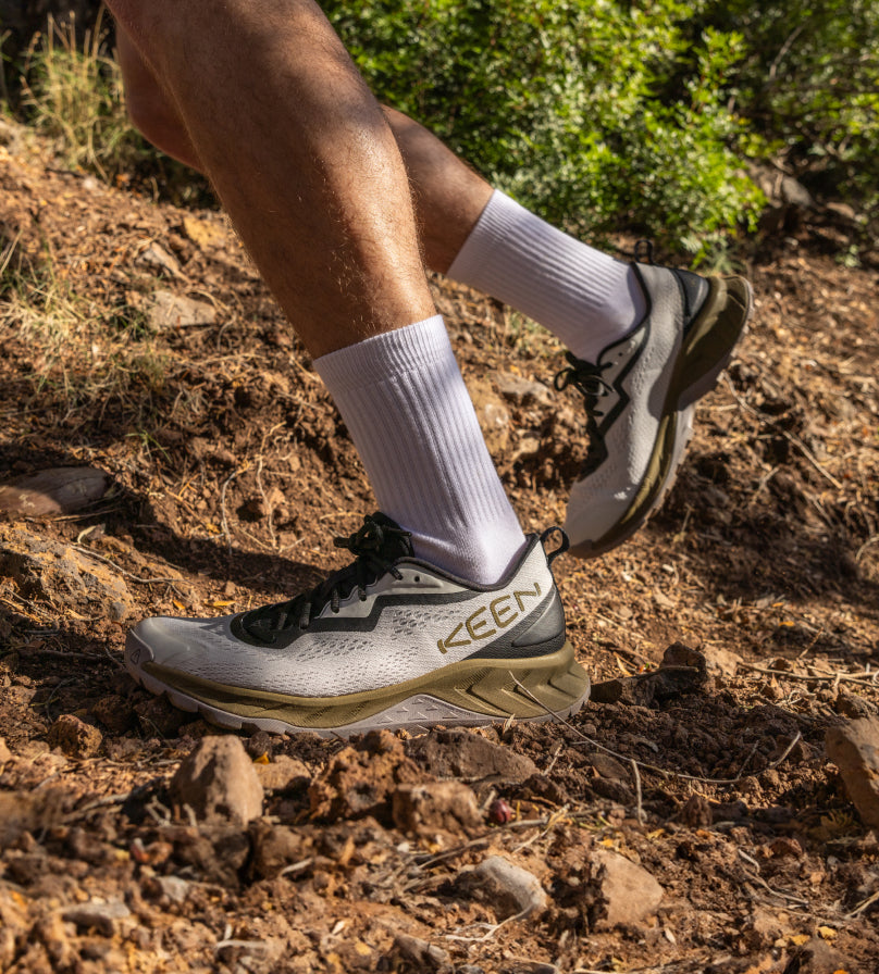 Knee-down shot of man wearing gray Versacore hiking shoe with gray crew socks while running on dirt path. 