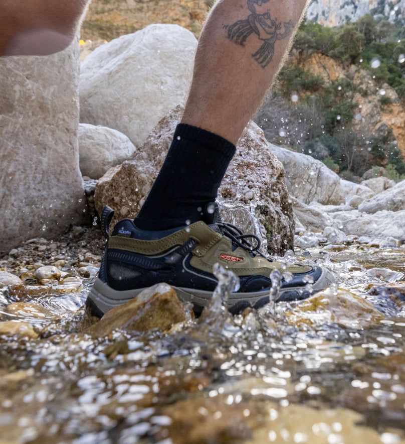 Knee-down shot of man walking through river stream while wearing olive green Targhee IV hiking shoes