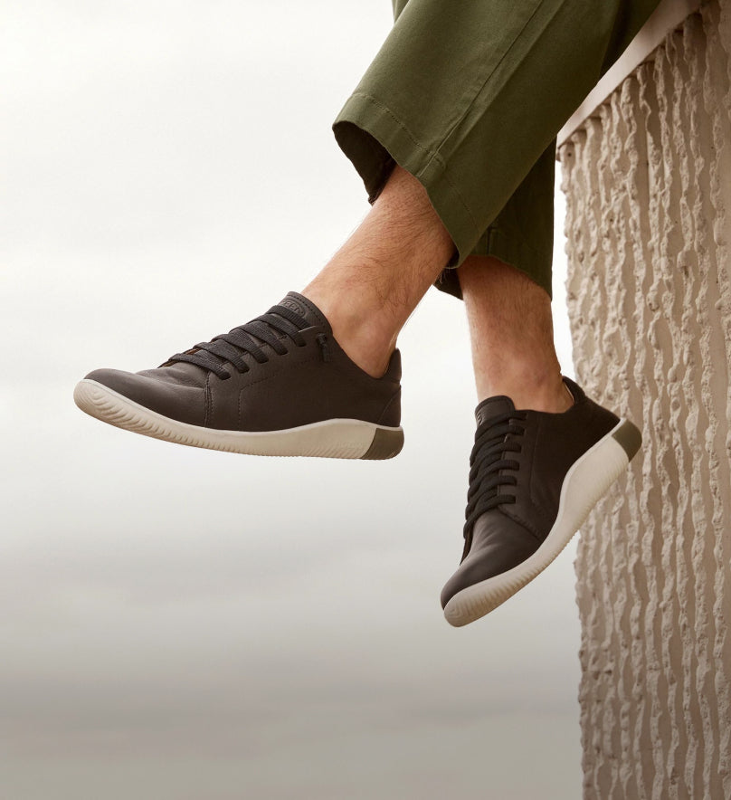 Knee-down shot of man sitting on cement ledge and dangling feet off while wearing black KNX sneakers 