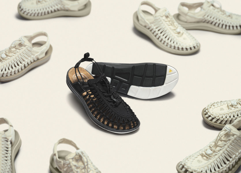Product shot of a black bair of the Mita and KEEN collab Uneek Sandals surrounded by other pairs of their white alternate color option. 