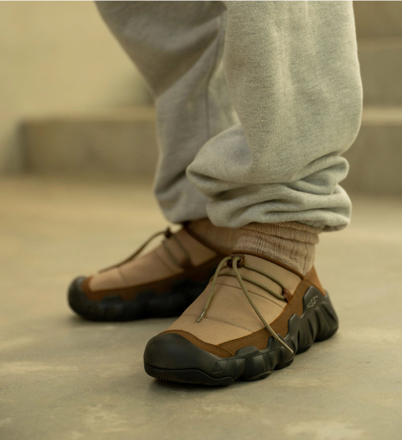 Knee-down shot of man in gray sweats wearing tan, brown, and black Hypowser shoes standing in front of cement stairs