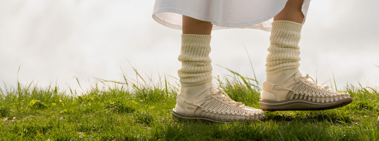 Knee-down shot of woman wearing tan socks and tan uneeks walking across grassy field. 