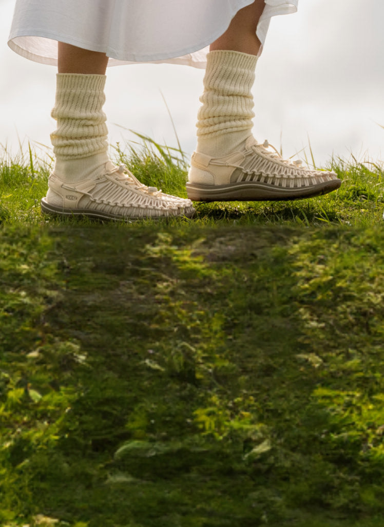 Knee-down shot of woman wearing tan socks and tan uneeks walking across grassy field. 