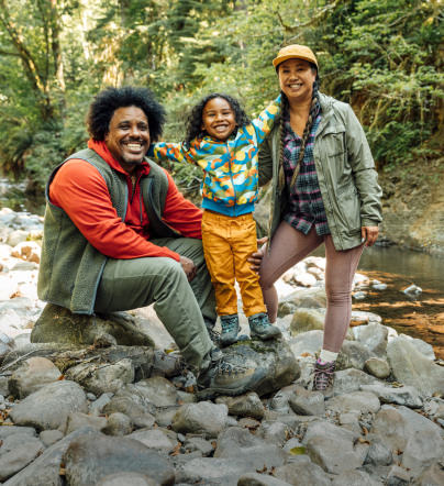 Family on rocky shore in front of river in hiking clothes and boots, posing for camera. 
