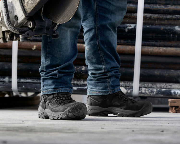 Knee down view of man wearing Pittsburgh energy workboots and jeans in front of metal pipes