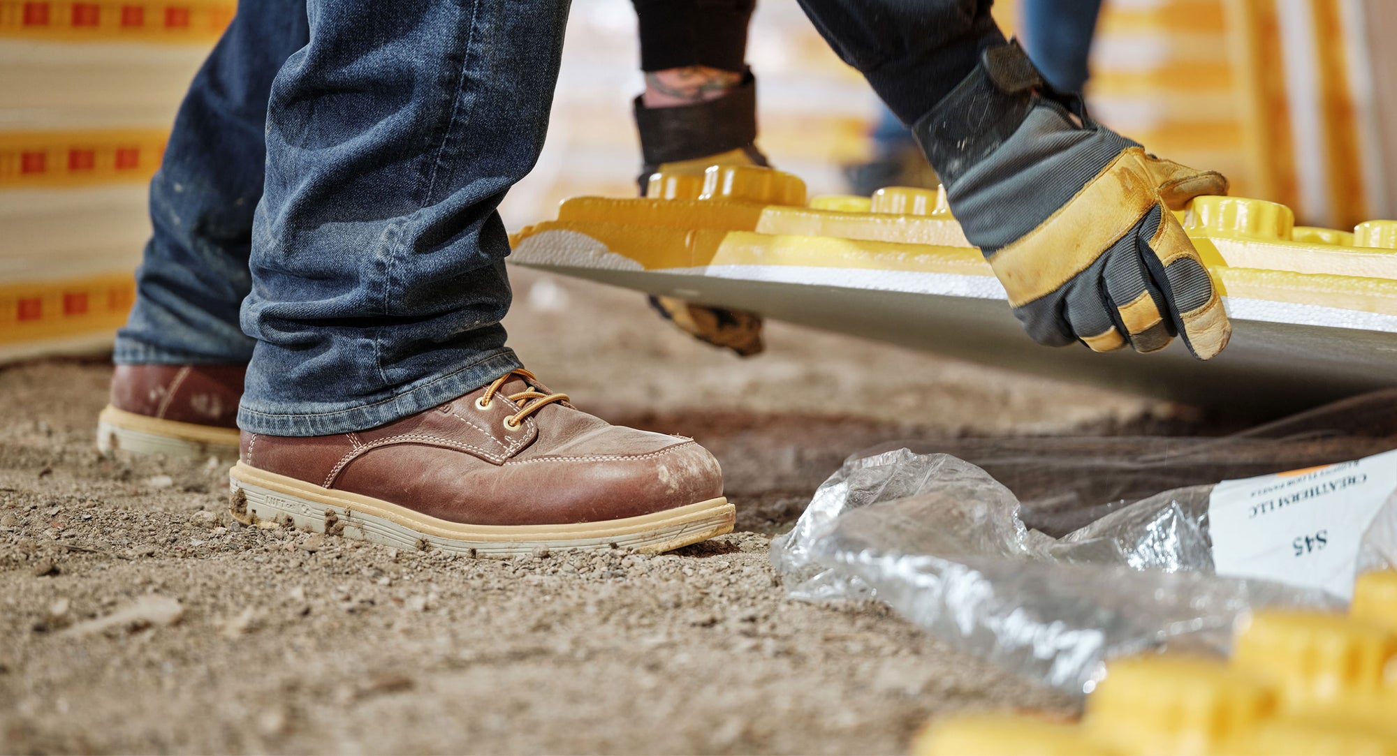Knee-down shot of man wearing San Jose work-boots and lifting building materials off of dirt ground 