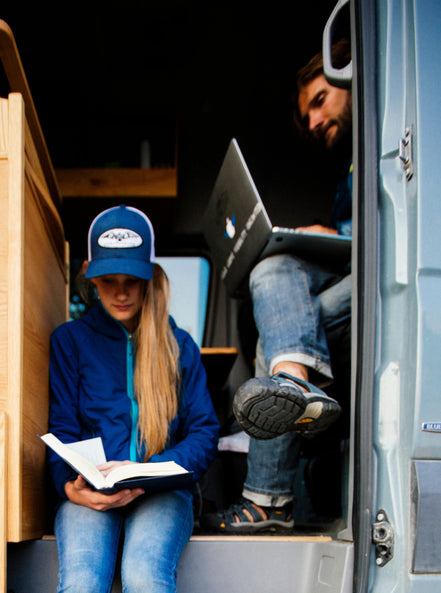 Man and Woman sitting in entry way of a built-out van reading and working on laptop