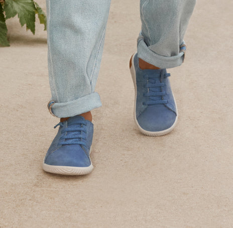 Knee-down shot of little boy in denim wearing blue leather KNX sneakers and walking on tan pavement.  