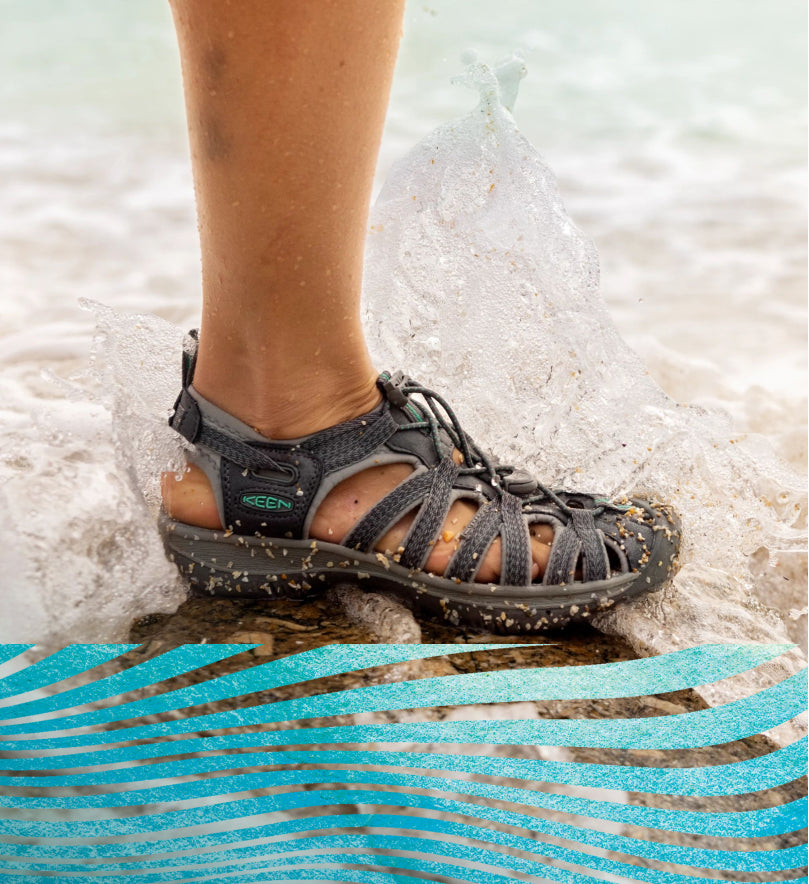 Knee down shot of woman wearing blue and grey Whisper sandals ad standing on rock in ocean water. 