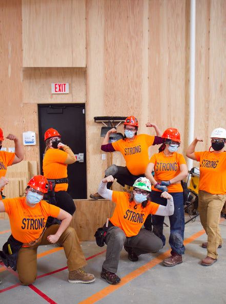 Tradeswomen posing in hard hats and orange shirts that say 