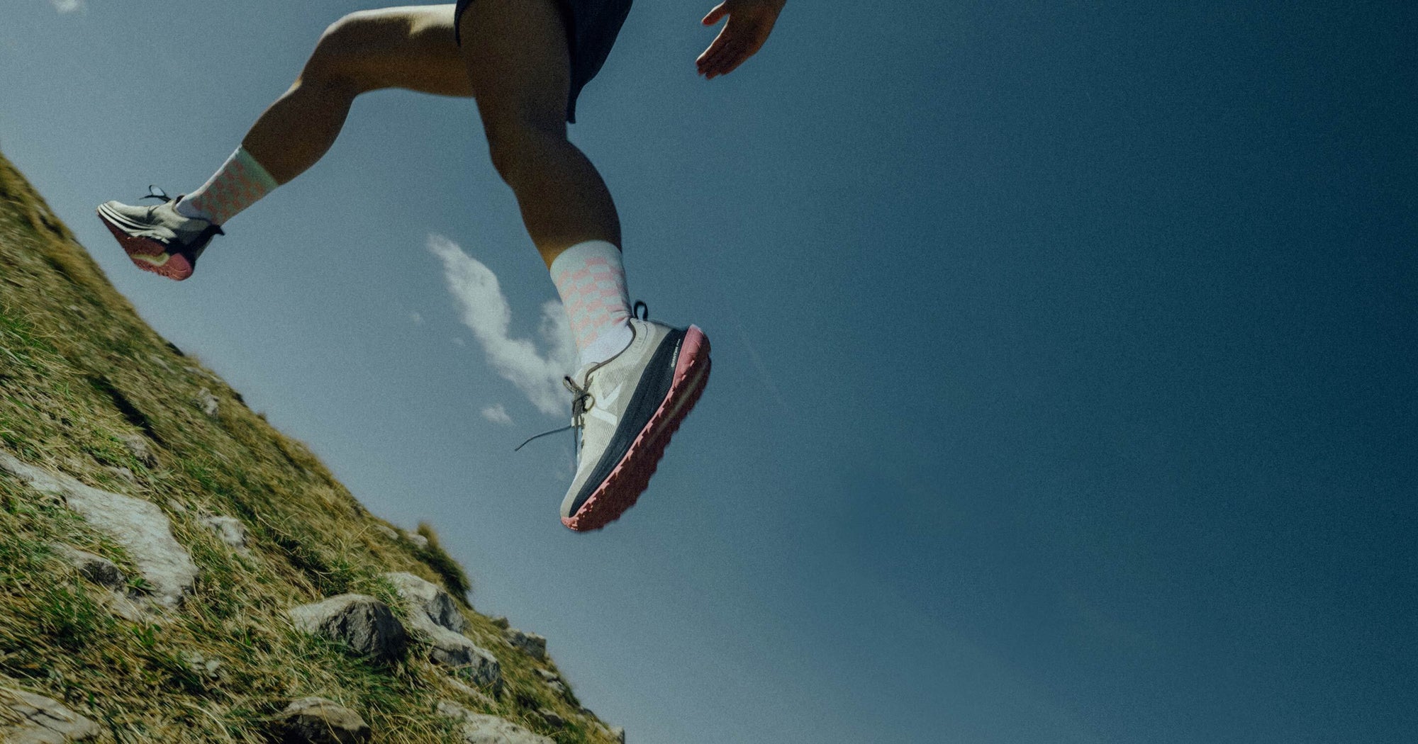 A woman wearing athletic shorts and blue and pink Roam running shoes in mid-stride  running across grassy plain. 