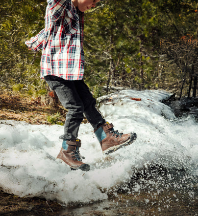 Waist-down shot of little kid wearing brown and blue Kanibou winter boots and kicking snow on winter trail 
