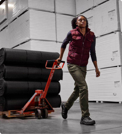 A woman in a warehouse, pulling large mats on a dolly.