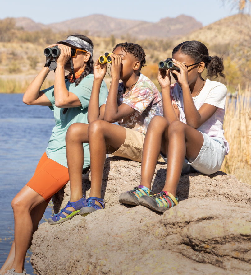 2 kids with an adult, all looking through binoculars across a lake and wearing newport h2 sandals