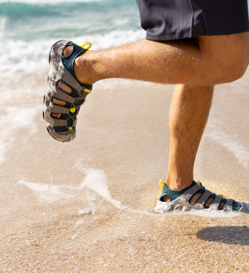 Knee-down shot of man wearing gray Hyperport sandals and running through wet sand on ocean shoreline. 