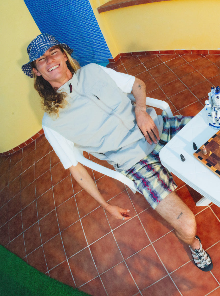 A man wearing plaid shorts and bucket hat playing chess on an orange tiled pool patio while wearing KEEN x Gramicci Newport sandals.