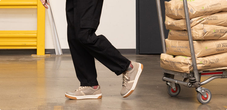 Knee-down shot of man wearing tan Kenton work sneakers and pulling a cart of brewing materials across warehouse floor. 