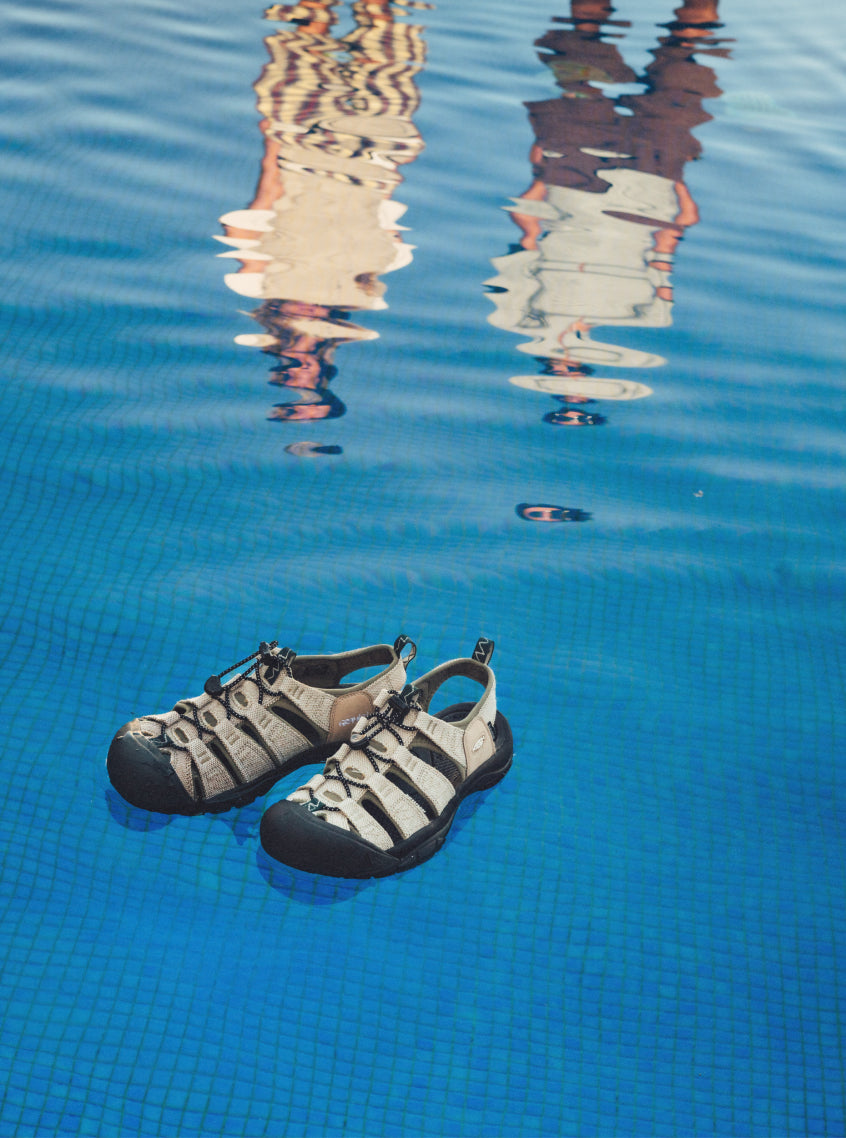 A pair of tan and black KEEN x Gramicci sandals floating on blue pool water with two peoples reflections near the edge.