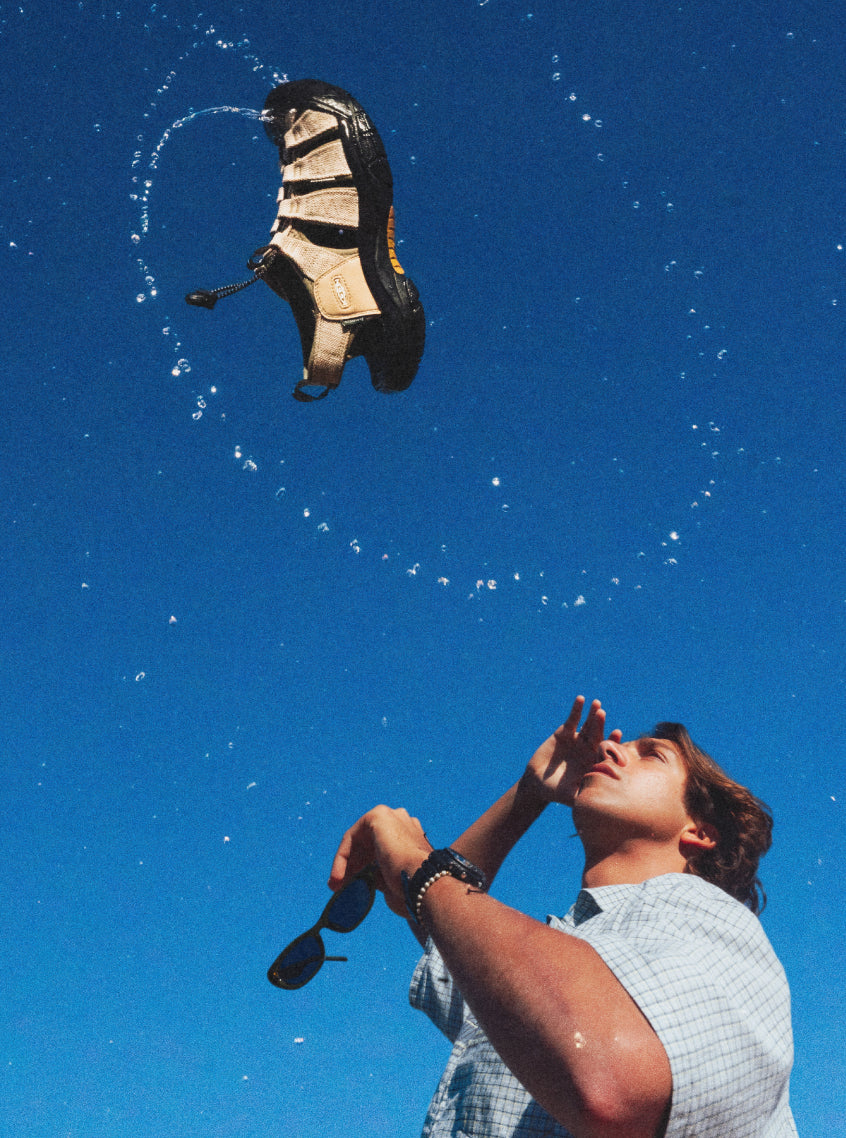 Stylistic shot of KEEN x Gramicci Newport Sandal being tossed into blue sky with water droplets spiraling around it.