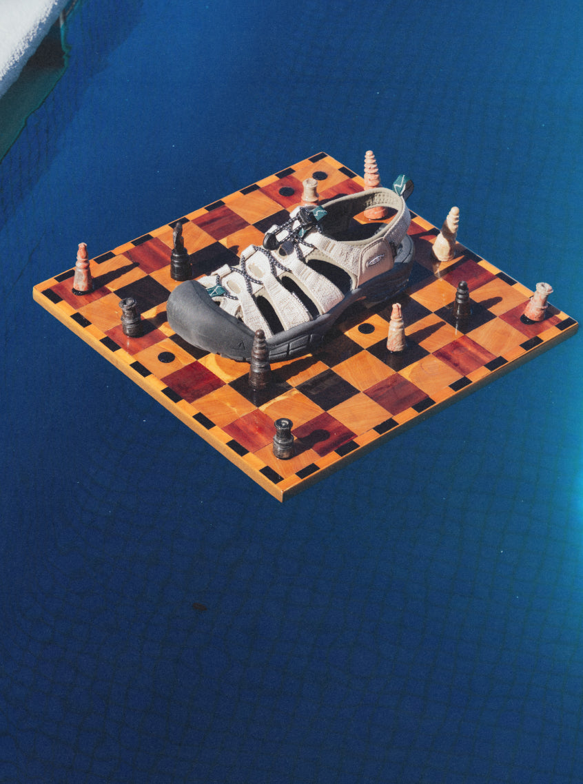 Solo KEEN x Gramicci  tan and black Newport sandal placed on wooden chessboard that is floating on blue pool water.